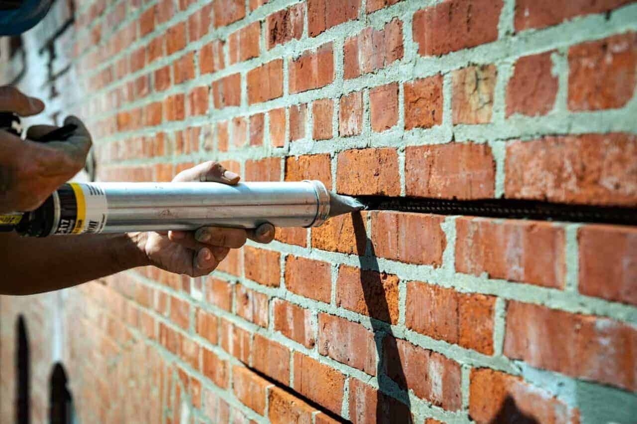 A person uses a caulking gun to fill a crack in a red brick wall.