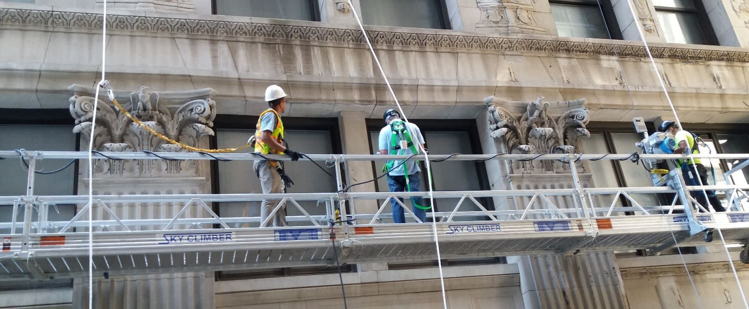 Workers in safety gear stand on scaffolding outside a stone building facade.