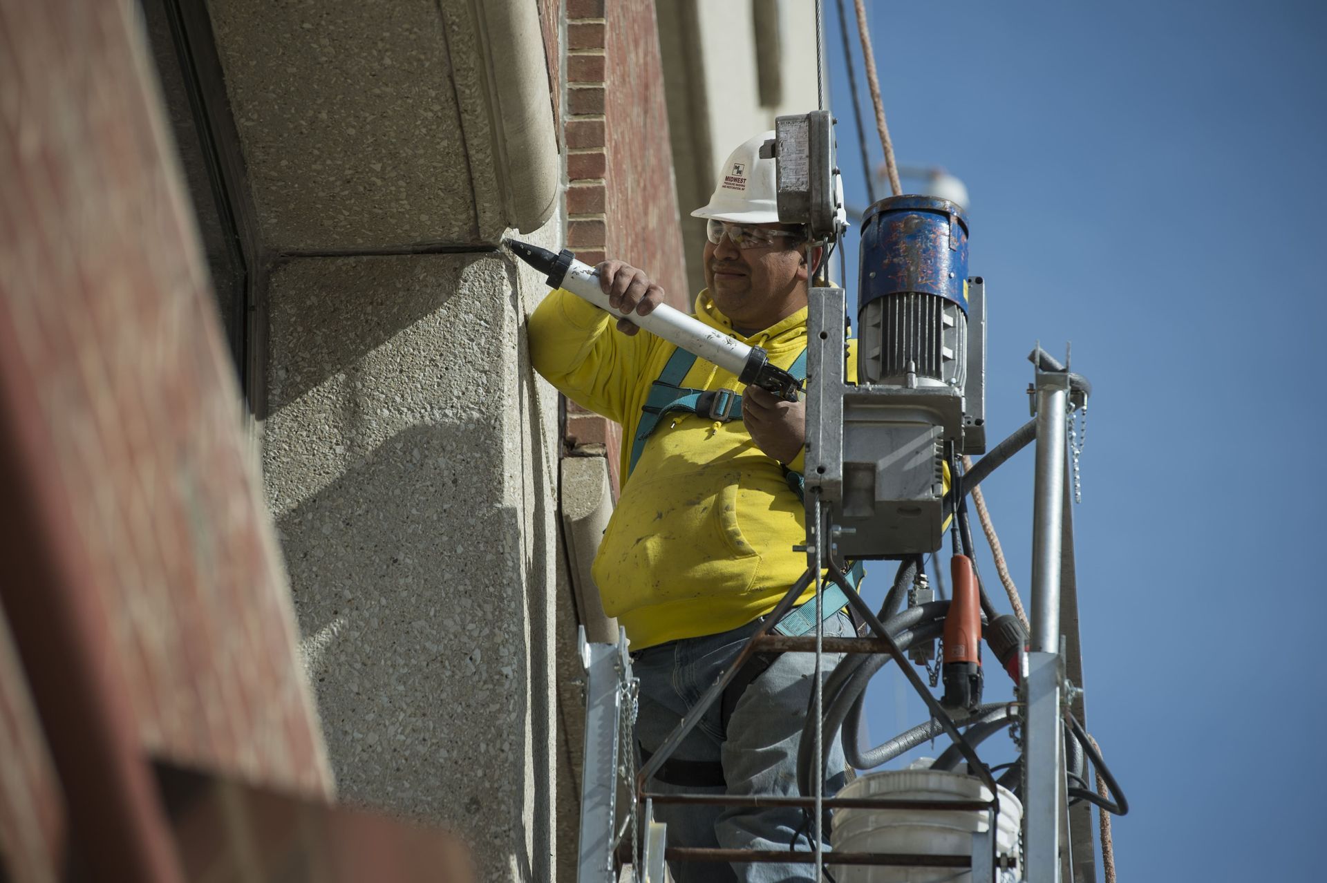 A worker in a yellow hoodie and safety harness on a suspended platform uses a caulking gun on a building exterior.