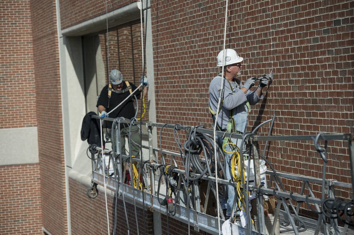 Two workers in hard hats use a suspended scaffold to repair bricks on the side of a tall brick building.