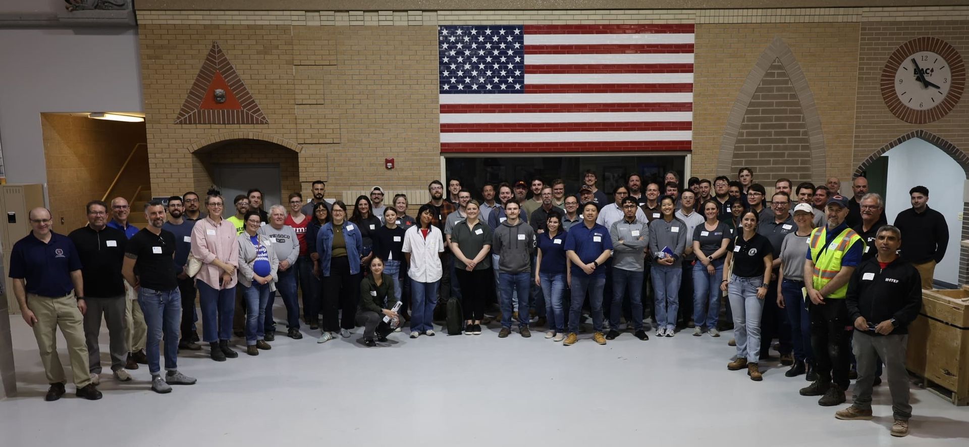 Large group posing indoors beneath a U.S. flag, with flags and a wall clock in the background.