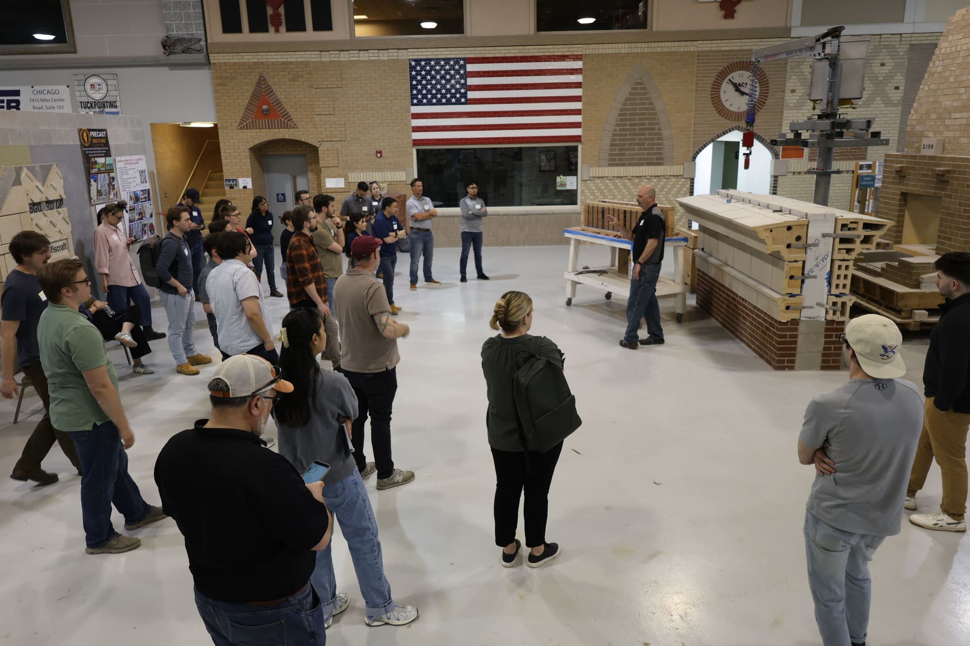 Group of people standing in a hall facing a speaker near a large American flag and display booths