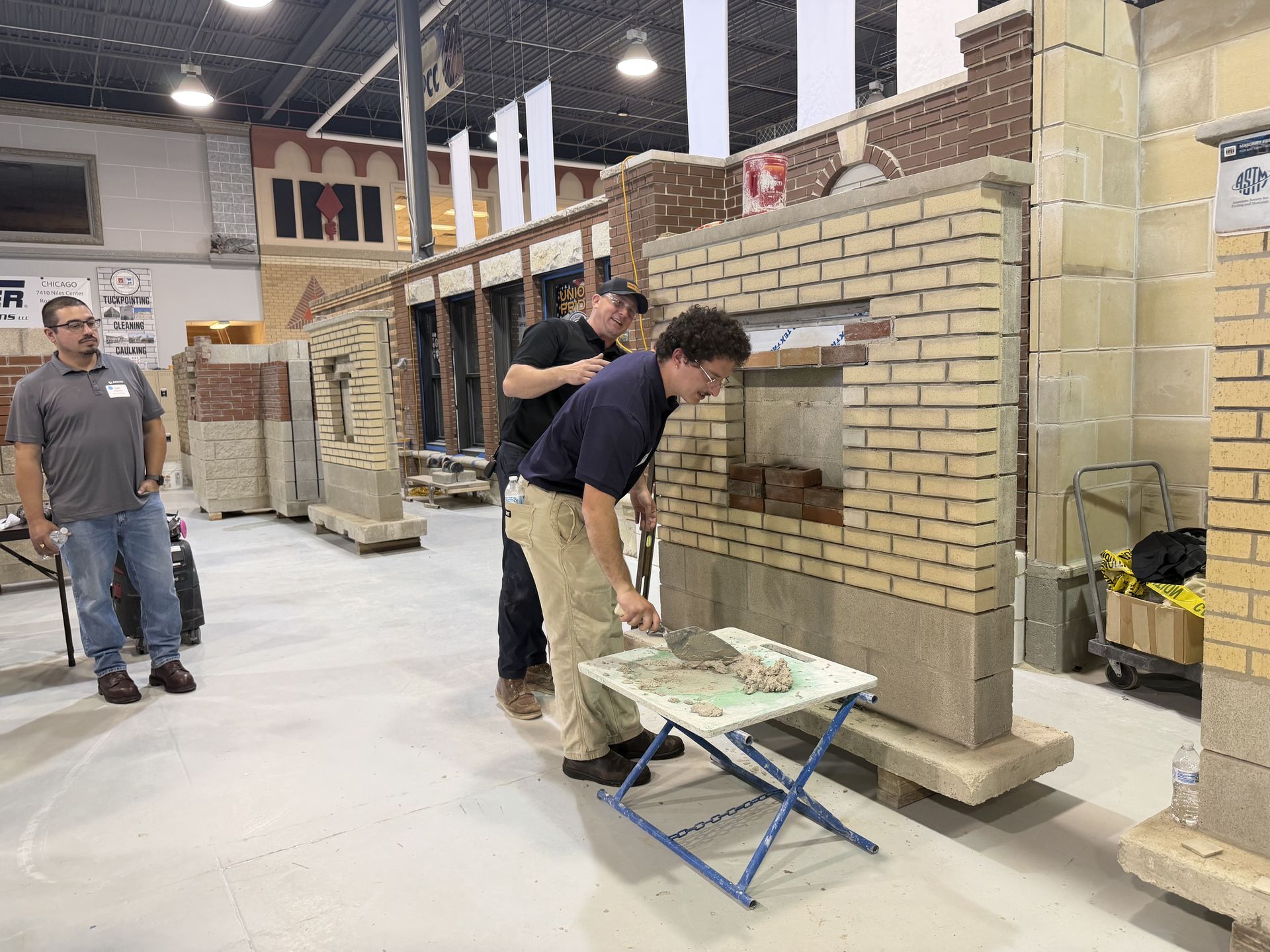 People building a brick wall in a workshop, with tools and a folding table in front.