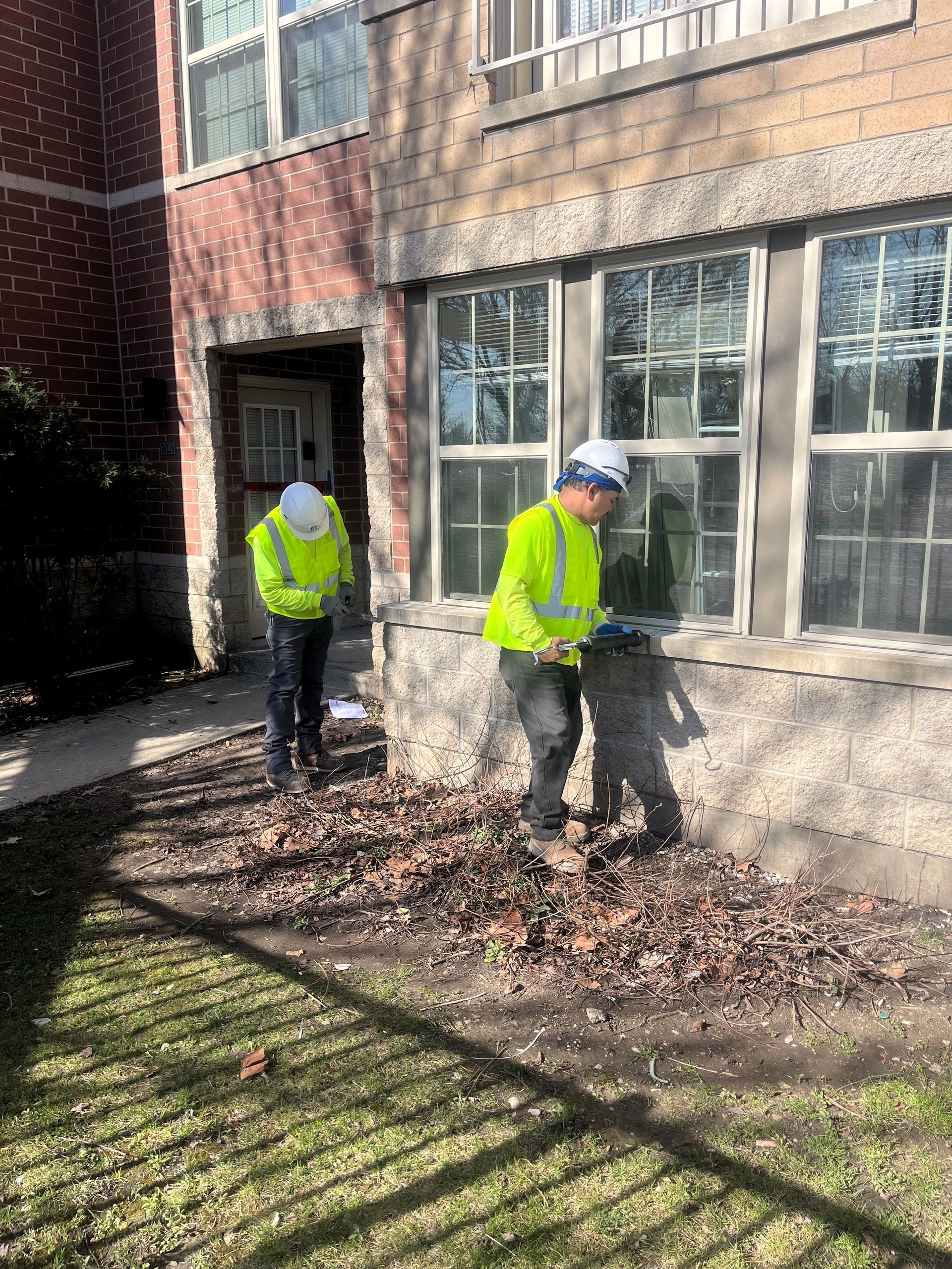 Two RestoreWorks employees wearing high-visibility jackets, hard hats, and gloves perform caulking and sealant services on an apartment complex in Chicago. One worker applies sealant along the window ledge using a caulking gun, while the other prepares materials nearby. The building features a mix of brick and stone masonry, and the team is working in a landscaped area with dried leaves and branches. The bright sunlight casts long shadows, emphasizing the attention to detail in the restoration process.