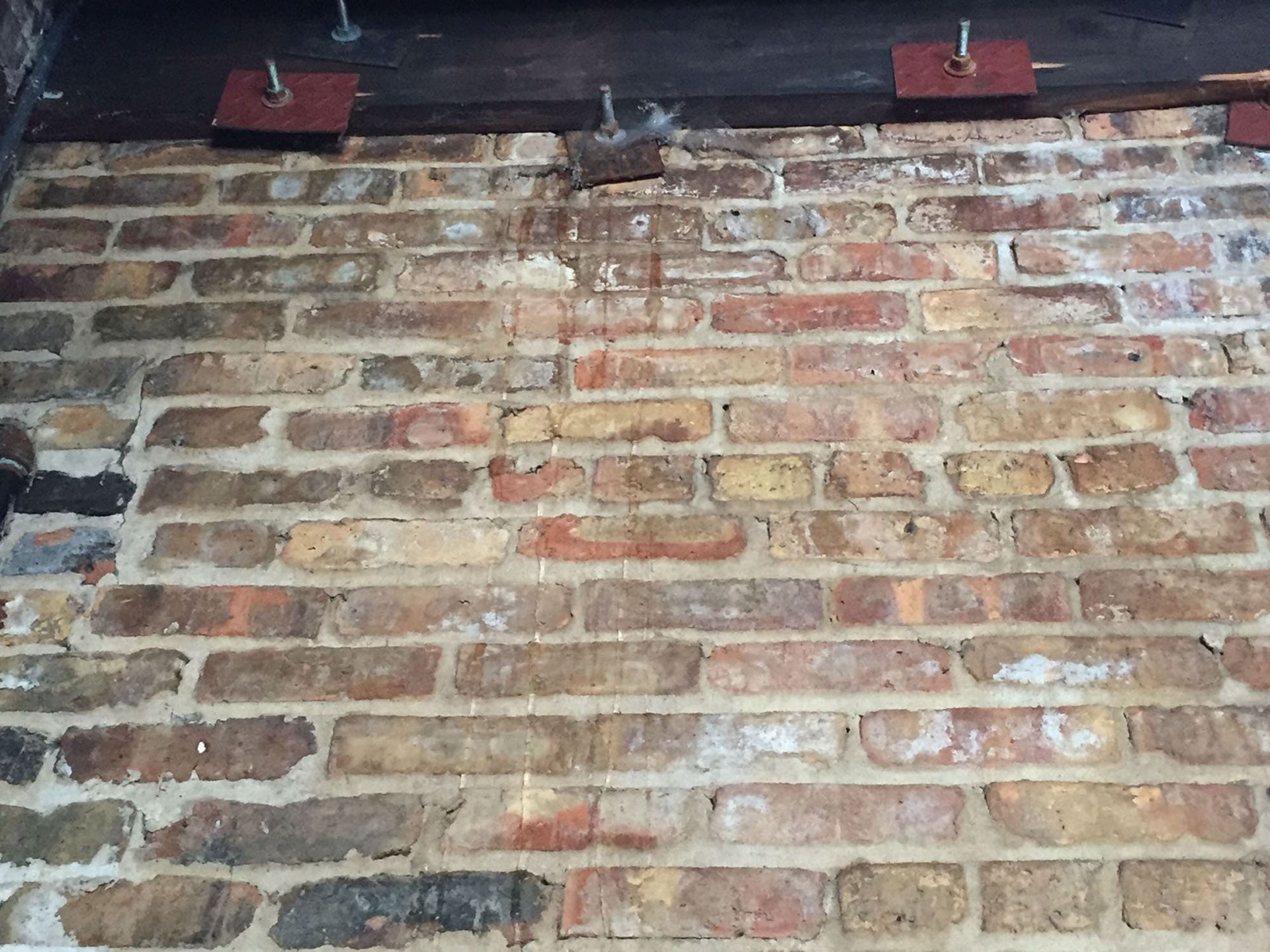 Close-up of a weathered brick wall with three red metal plates bolted to a wooden beam along the top edge.
