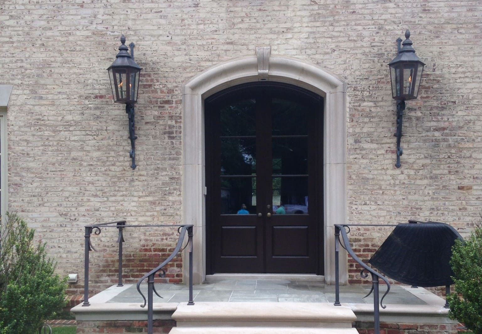 A dark double door with a stone archway, framed by two vintage-style lanterns on a light-colored brick exterior.