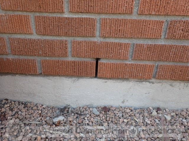 Brick wall with a vertical crack above a concrete foundation and gravel ground below
