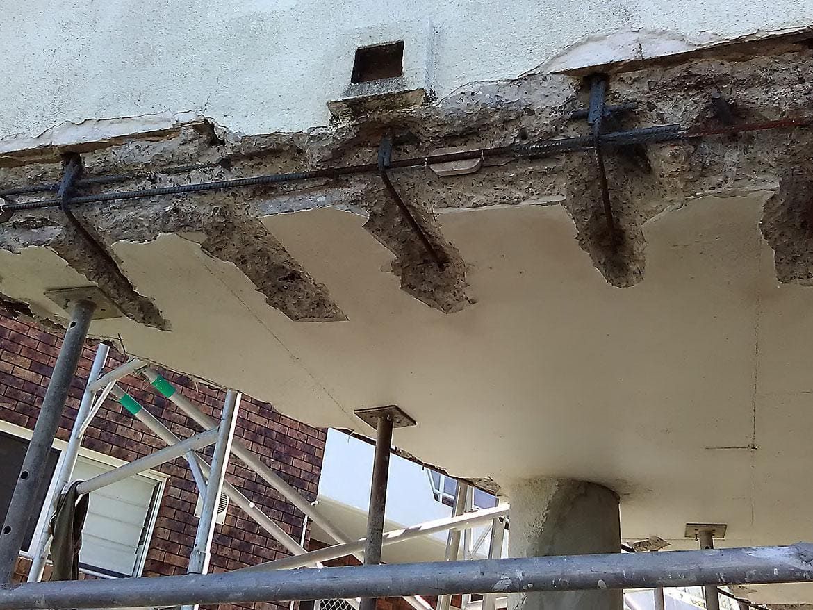 Damaged concrete balcony underside with exposed rebar and support posts during construction repairs