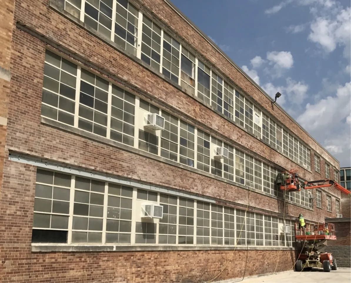 A red brick multi-story building features rows of large windows and a worker on a lift servicing the facade.