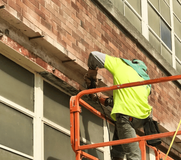 Worker in neon safety shirt cleaning a brick building from an orange lift platform.
