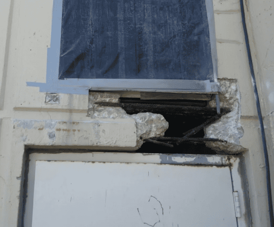 Damaged exterior wall with a broken opening, cracked plaster, and taped window above