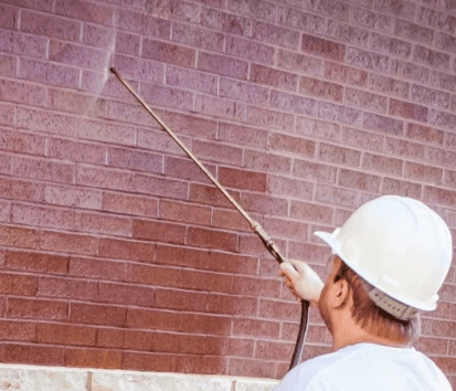 Worker in a white hard hat power-washing a brick wall with a long sprayer.