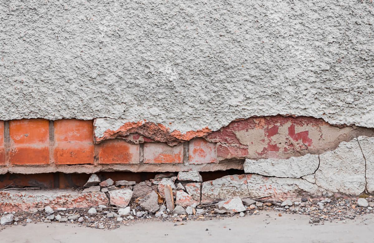 Close-up of a deteriorating masonry wall with severe spalling, exposing red brick beneath the cracked and crumbling stucco layer. Loose debris and broken mortar are visible at the base, indicating structural damage likely caused by moisture infiltration and freeze-thaw cycles.