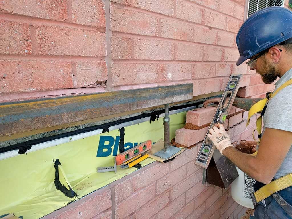 Worker installing insulation beneath a brick wall, using a power tool and ladder.