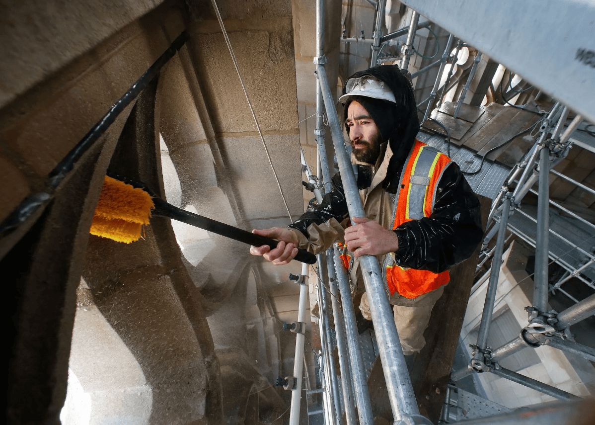 Worker in orange safety vest cleaning a metal structure with a long-handled brush in an industrial setting