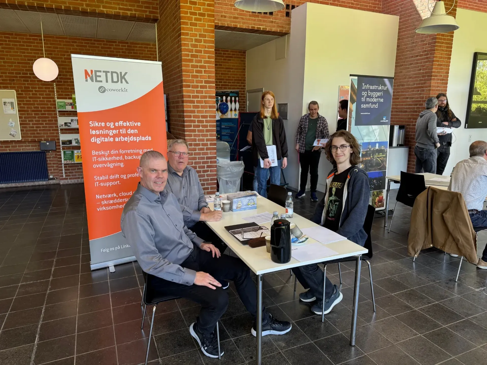 Folk ved en stand, der promoverer NETDK-tjenester. To mænd sidder ved et bord, den ene med en bærbar computer.
