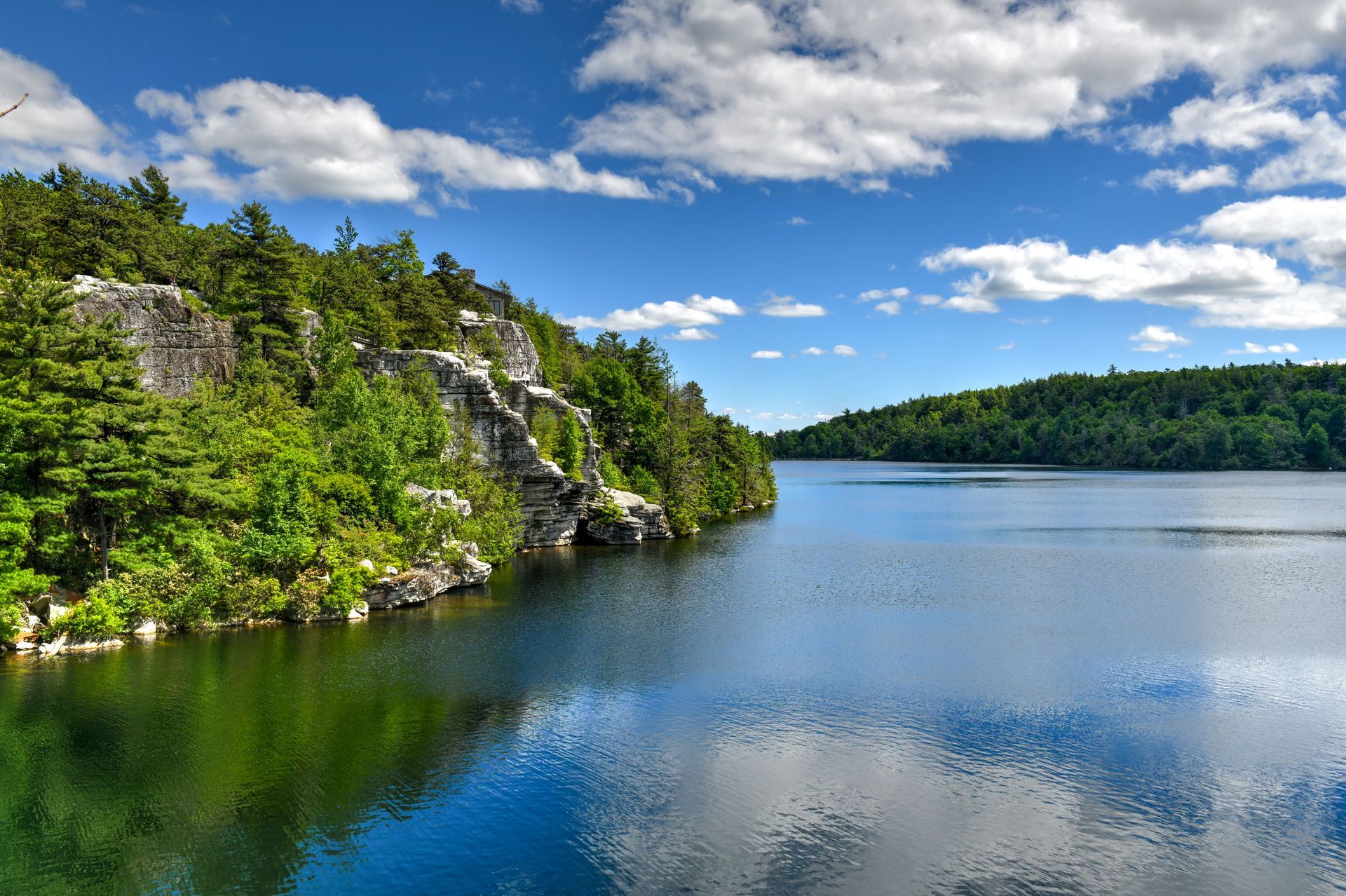Rocky green shoreline beside a calm blue lake under a bright, cloudy sky