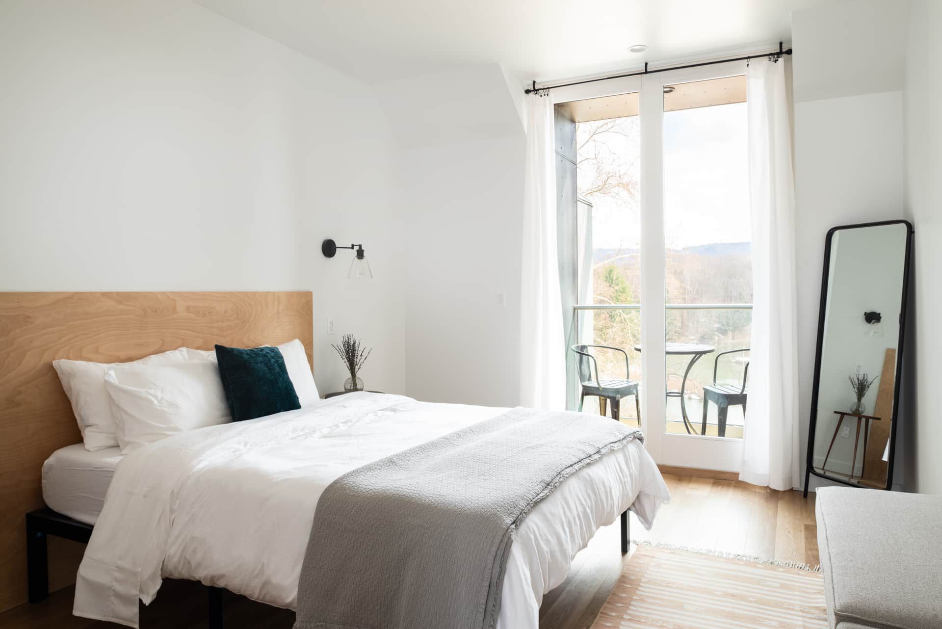 Bright bedroom with a bed, floor mirror, and open balcony doors letting in sunlight