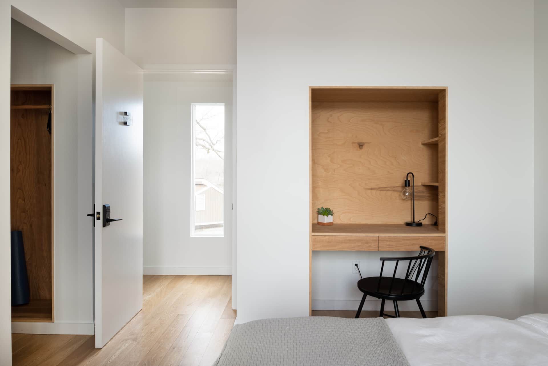 Minimal bedroom with a built-in desk nook, white walls, wood floors, and an open doorway to a bright hall