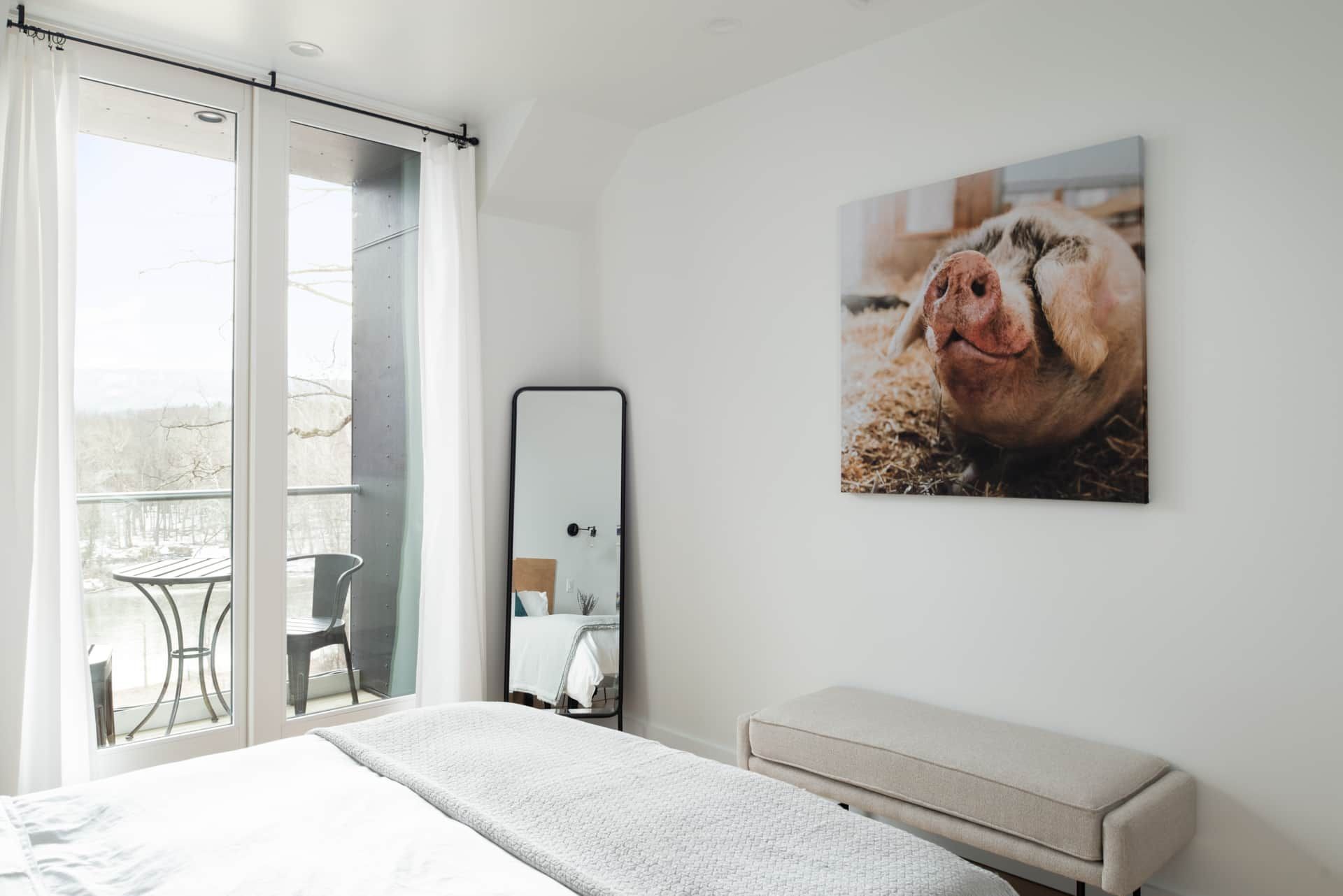 Bright bedroom with a pig portrait, mirror, bench, and open balcony doors