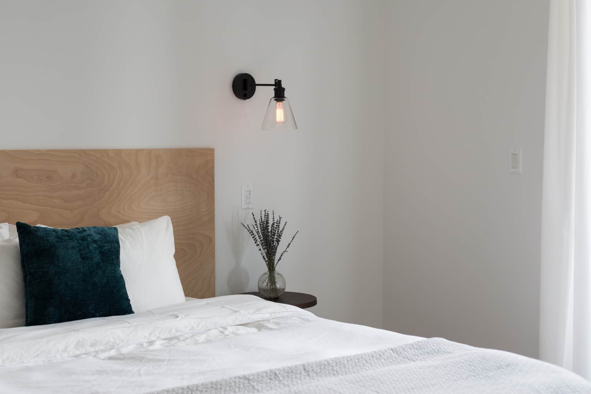 Minimalist bedroom with a bed, wood headboard, teal pillow, wall lamp, and white curtains