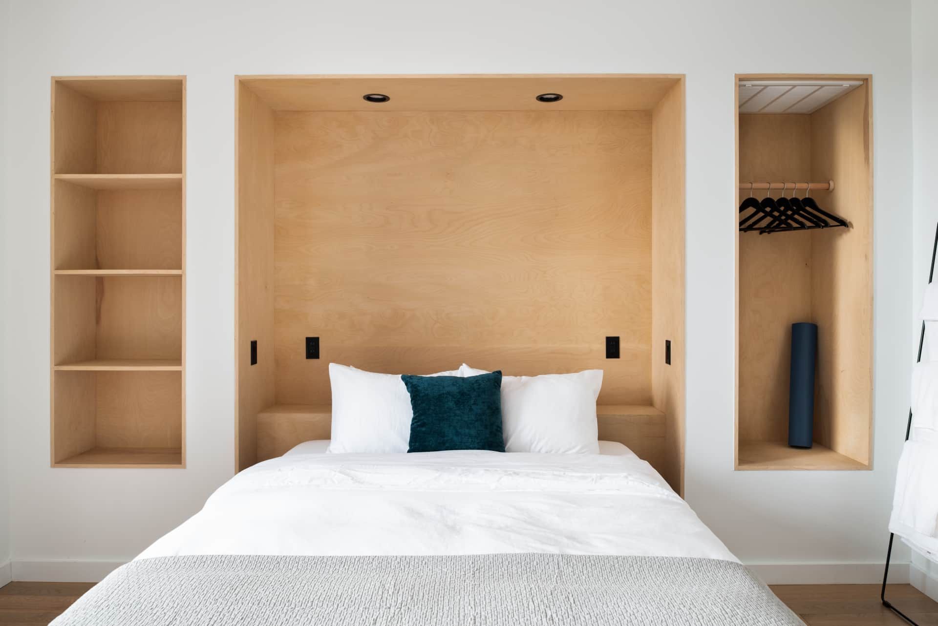 Minimalist bedroom with built-in wooden shelving, a bed, and a closet niche with hanging clothes and a blue vase
