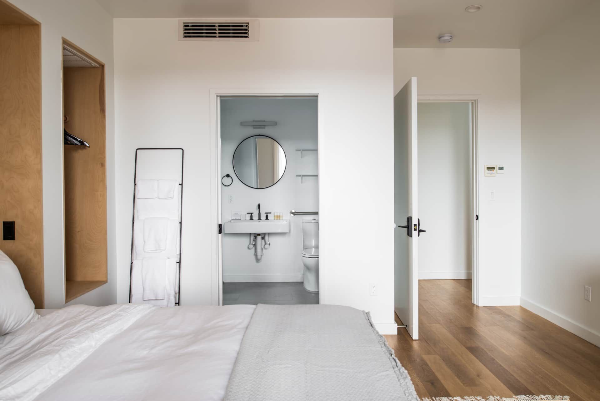 Bright bedroom with a bed, wood floor, and an open bathroom sink area with a round mirror.