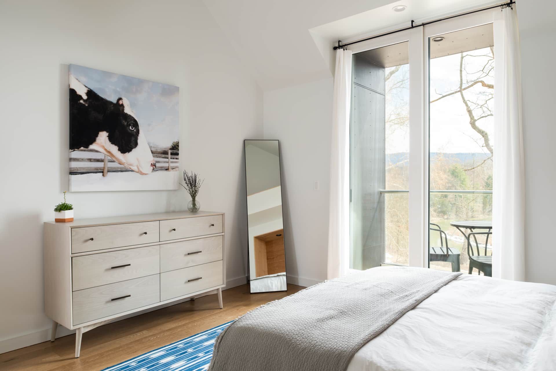 Bright bedroom with white dresser, leaning mirror, blue rug, and open French doors to a balcony.