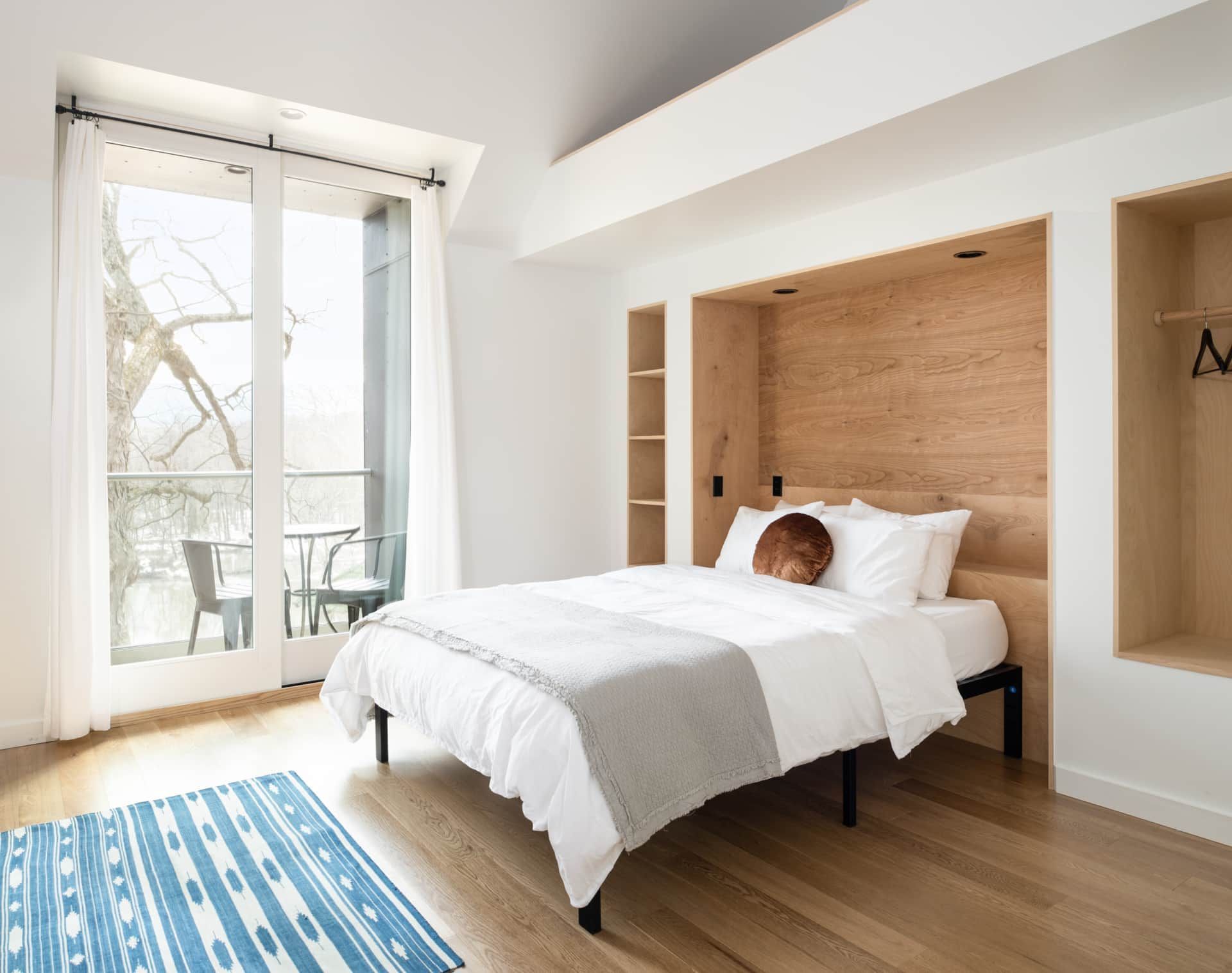 Bright bedroom with white bed, wood headboard, blue rug, and a glass door to a balcony.