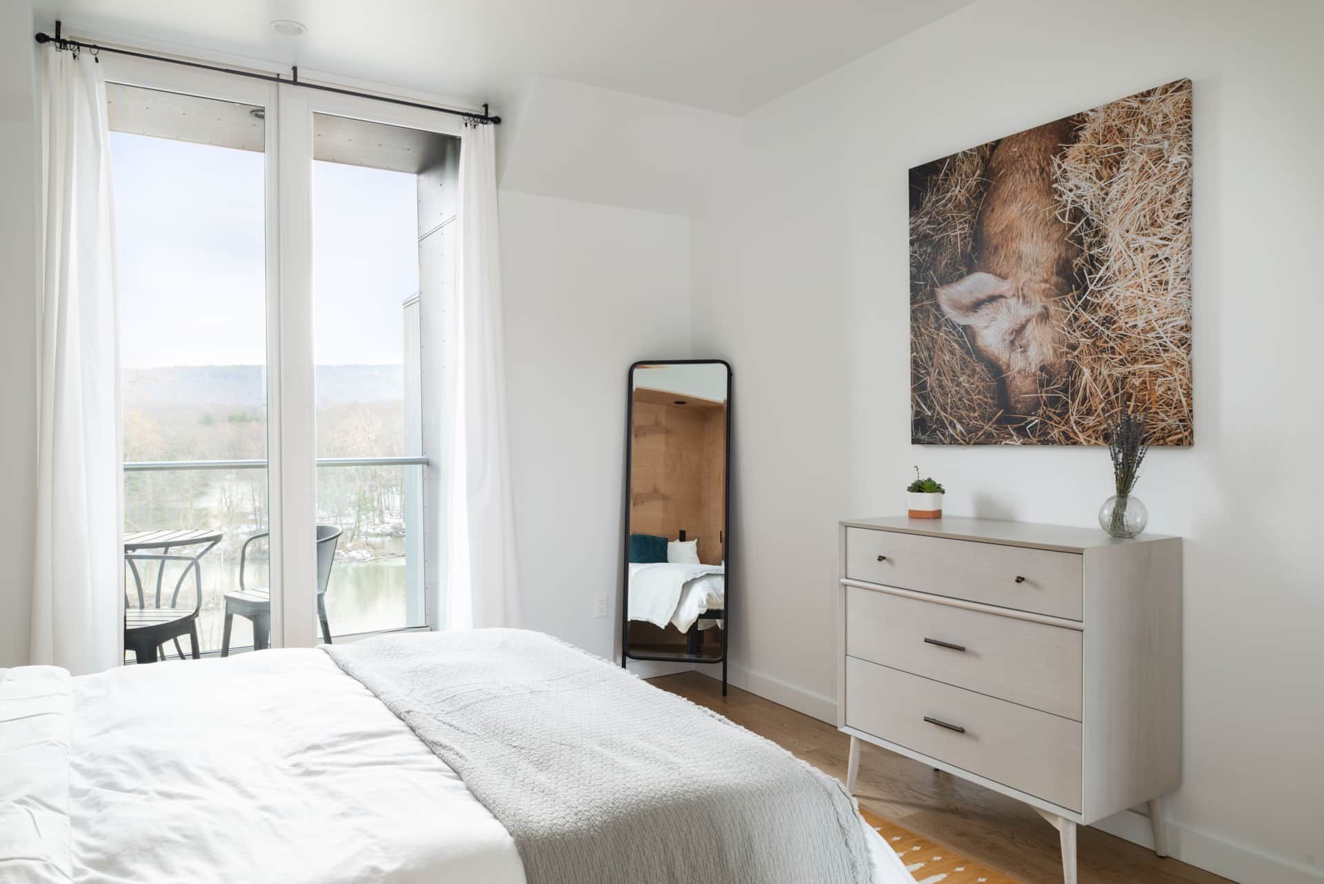 Bright bedroom with white bed, dresser, floor mirror, and balcony doors; lion artwork on the wall