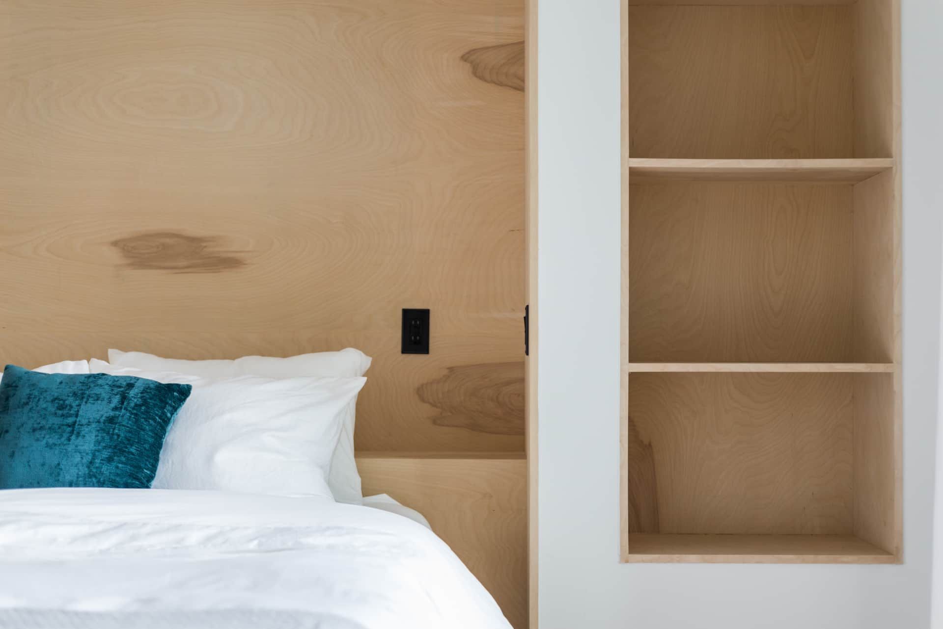 Bed with white linens and teal pillow beside built-in wooden shelf in a minimalist room