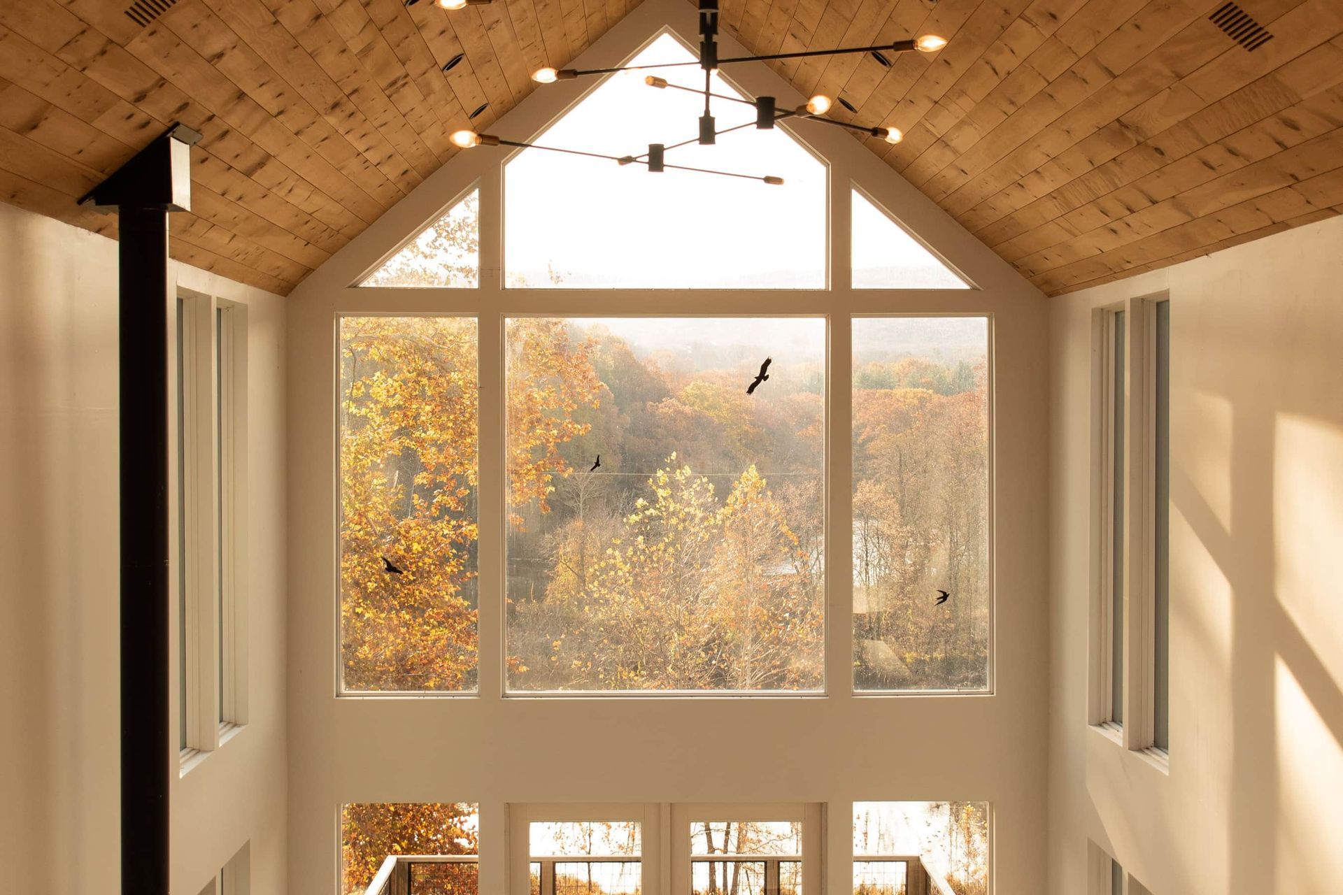 A bright room with a high wood ceiling, large triangular window, and autumn trees outside.