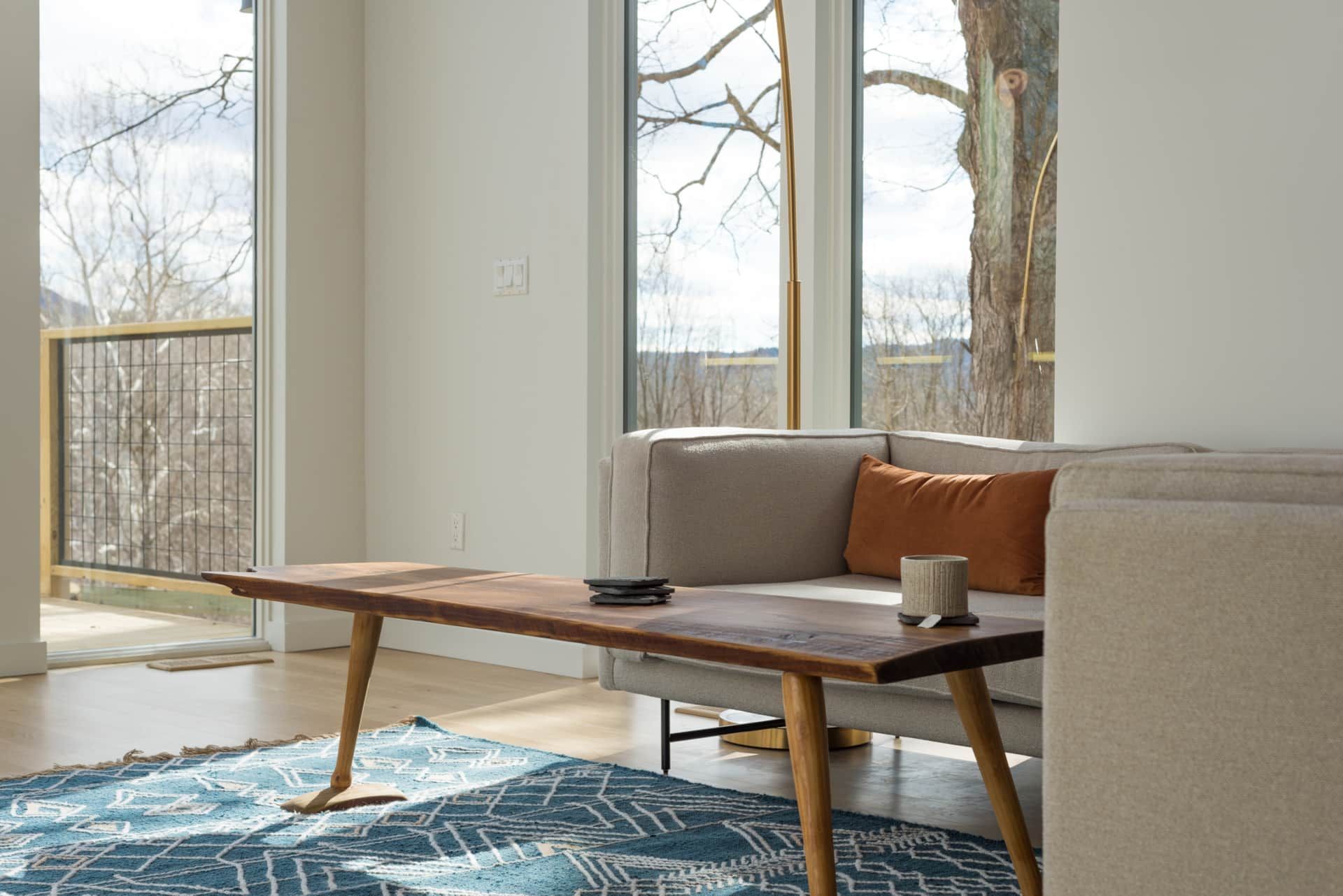 Modern living room with a beige sofa, wooden coffee table, blue rug, and large windows overlooking trees