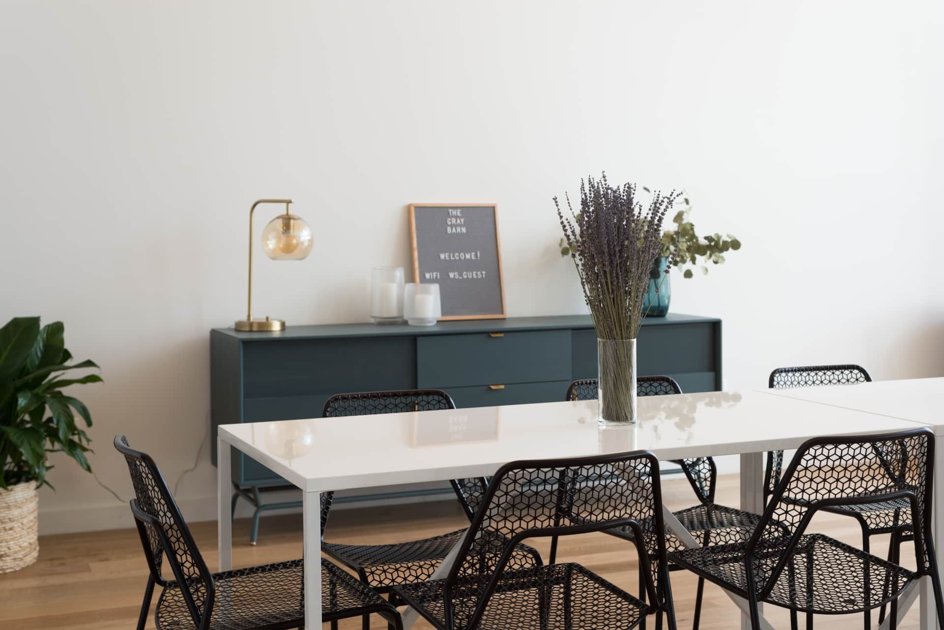 Minimal dining room with white table, black chairs, dark sideboard, lamp, and dried flowers.