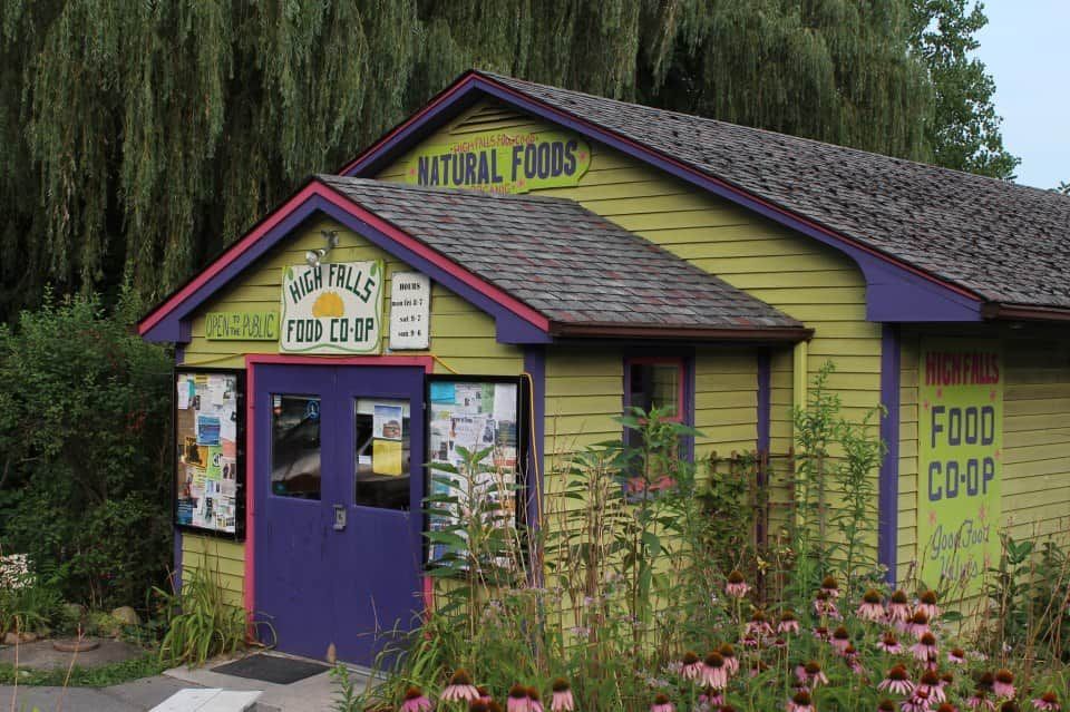 Purple and yellow natural foods co-op storefront with flower garden and green trees.
