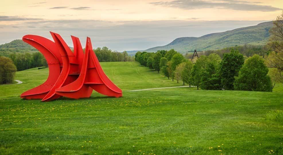 Large red abstract sculpture on a green lawn with hills and trees in the background at sunset