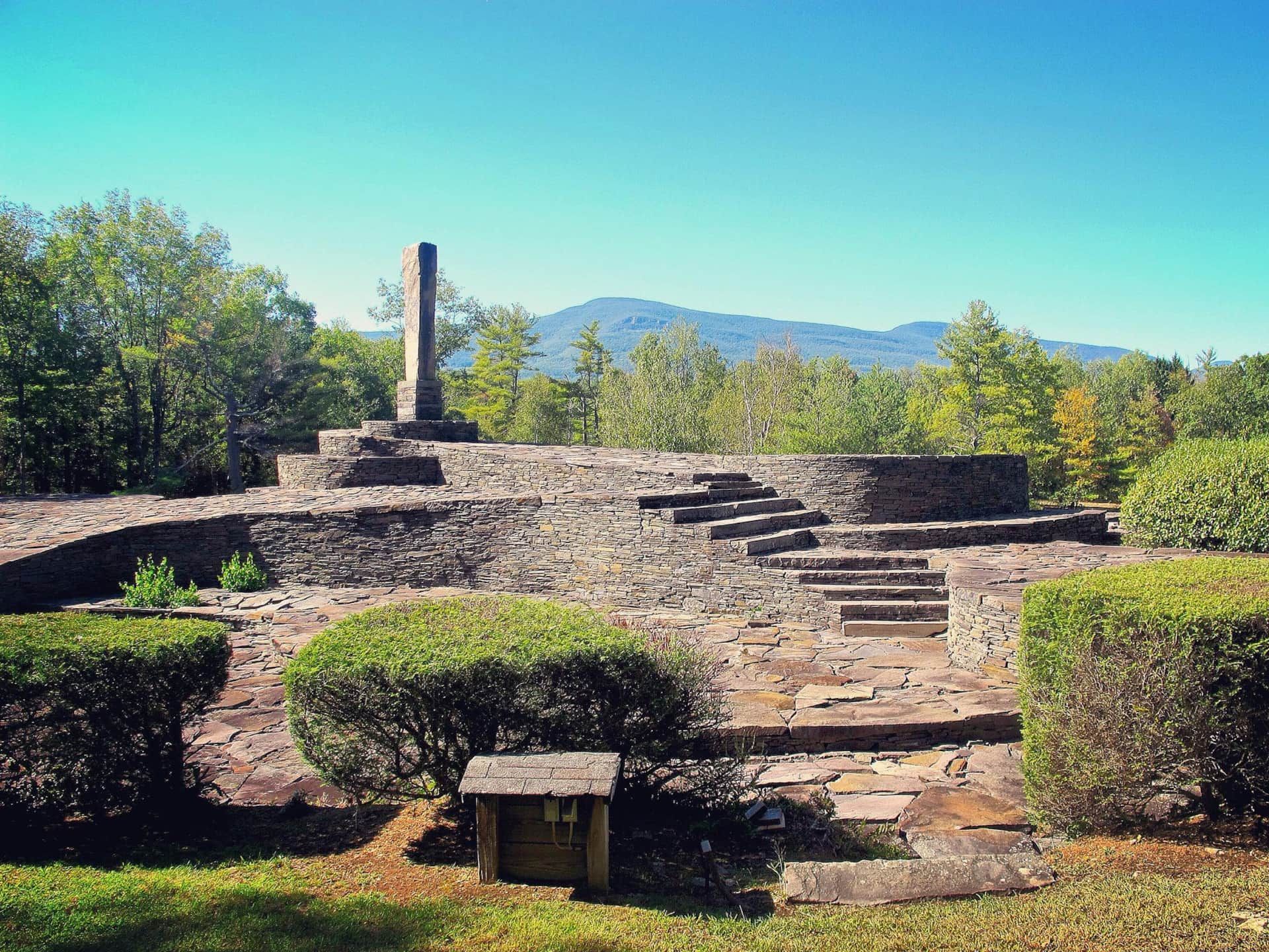 Stone ruins with steps and trimmed shrubs on a hillside, with mountains and blue sky in the background
