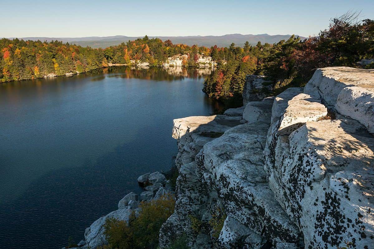 Rocky lakeside cliff overlooking calm blue water and autumn trees under a clear sky
