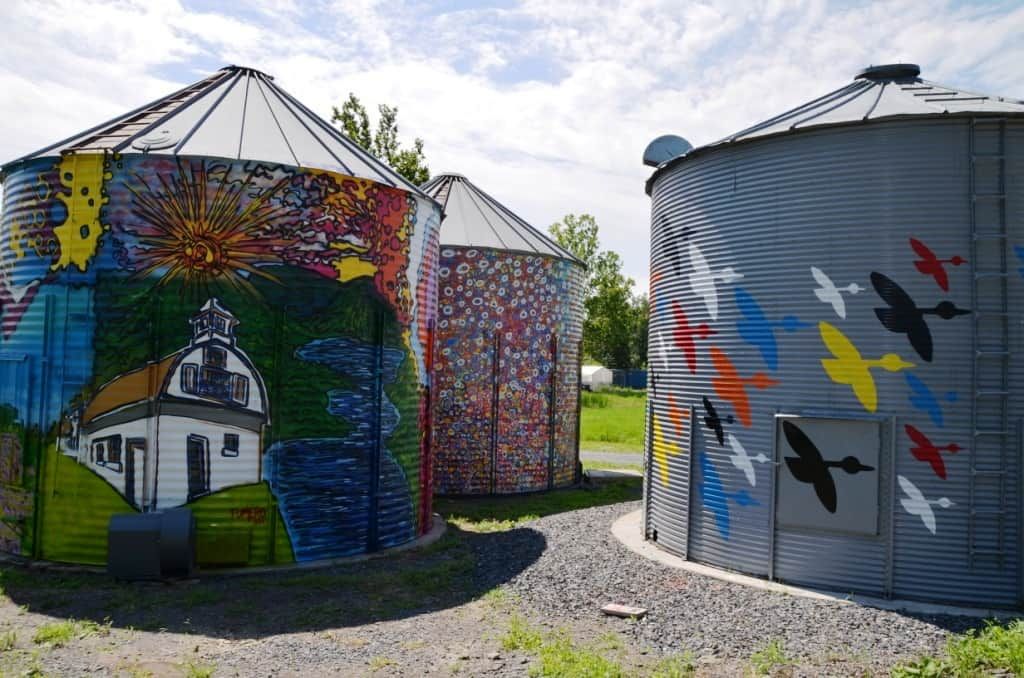 Colorful mural-painted grain silos in a gravel lot under a partly cloudy sky.