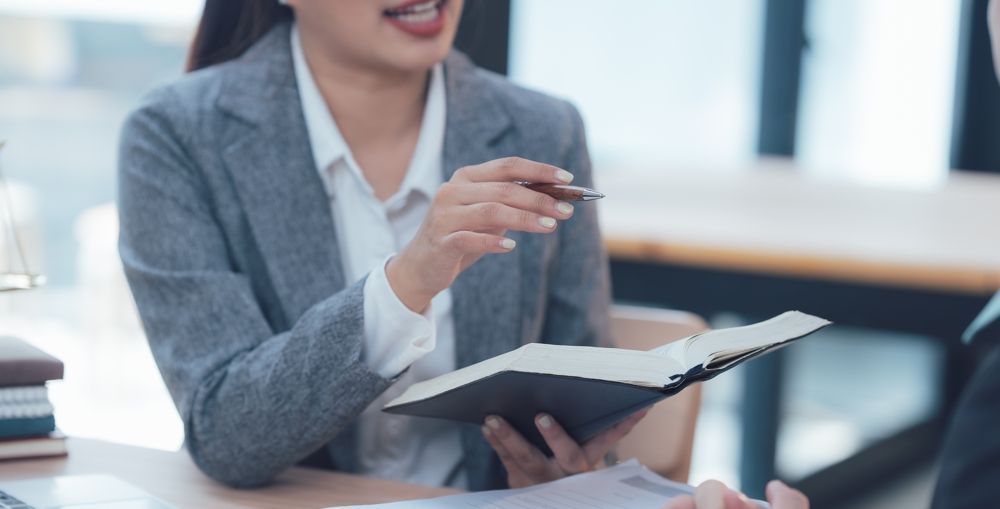 Woman in gray blazer gestures while holding open book, discussing with another person at a desk.