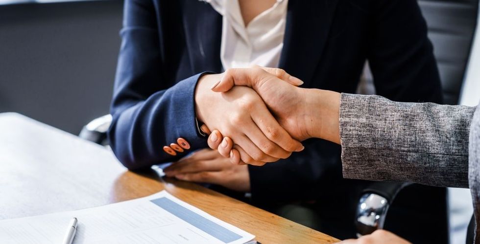 Two businesswomen shaking hands at a wooden desk, wearing professional attire.