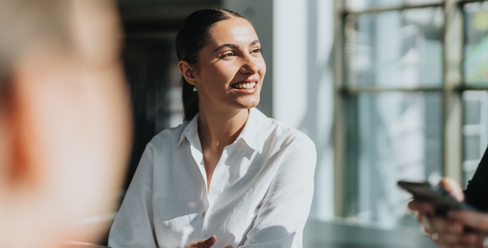 Woman in white shirt smiles, looking towards light, in an office setting.