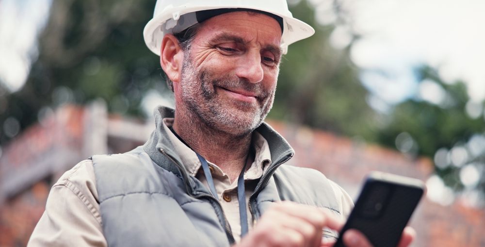 Construction worker in white hard hat smiles while using a smartphone outdoors.