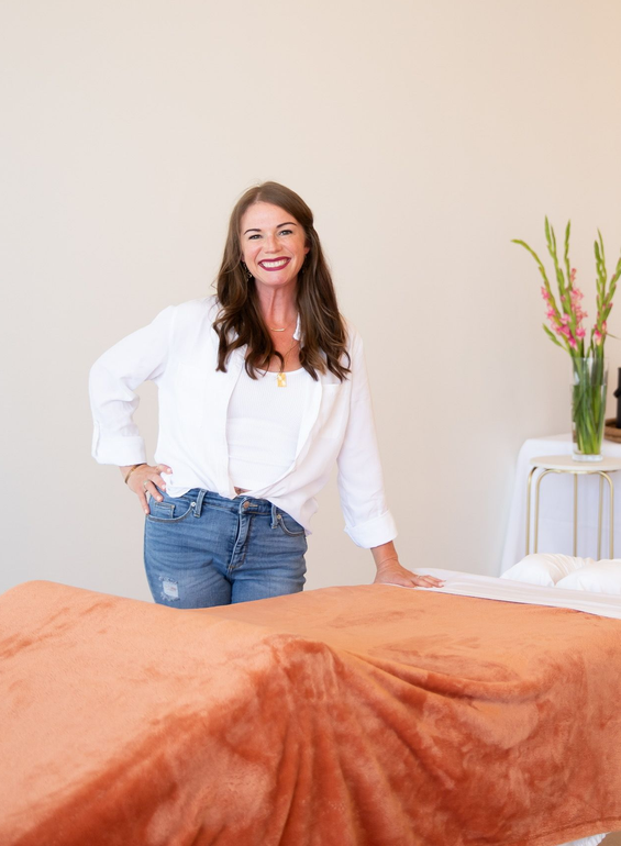 Woman in a hat and orange shirt smiling, sitting indoors against patterned wallpaper.