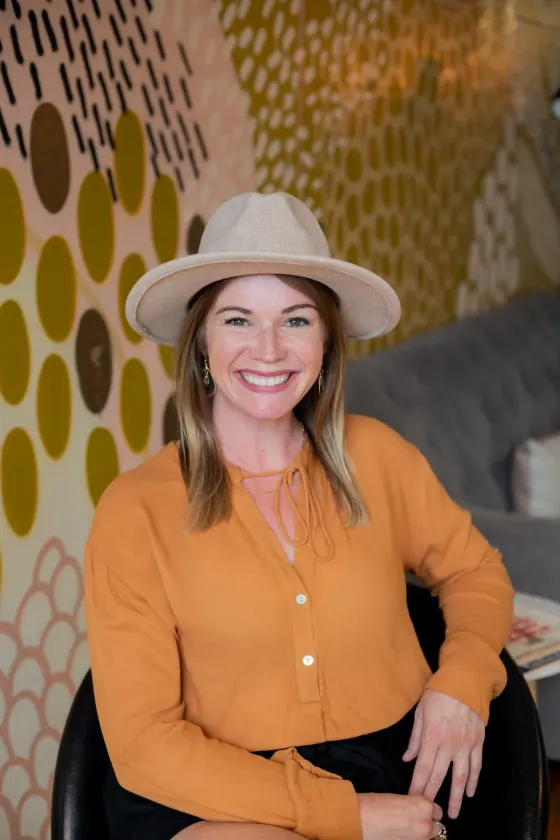 Woman in a hat and orange shirt smiling, sitting indoors against patterned wallpaper.