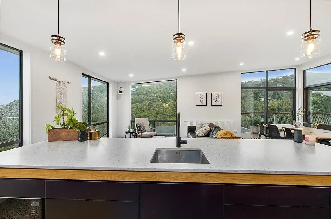 Modern open kitchen with white island, pendant lights, large windows, and a living area beyond