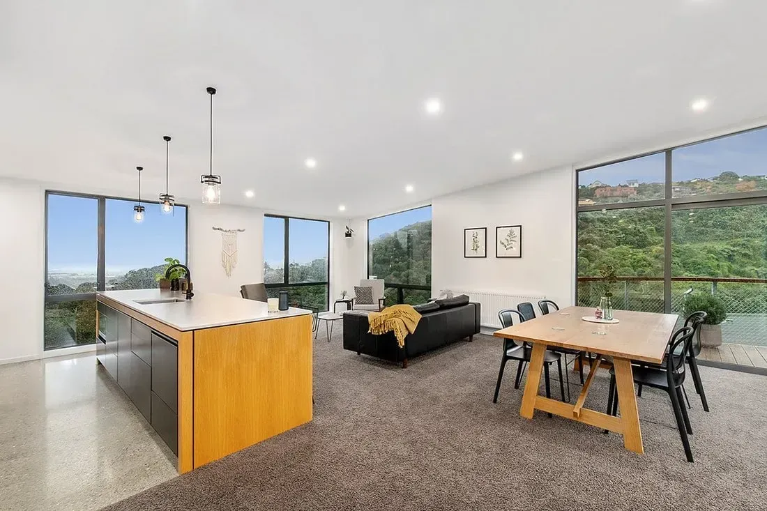 Open-plan kitchen and dining room with island, black sofa, and large windows overlooking greenery