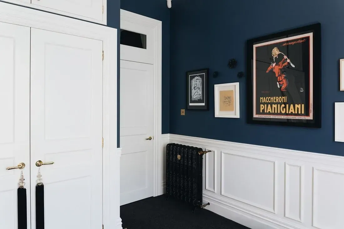 Blue-walled room with white paneled doors, black radiator, and framed artwork above chair rail.