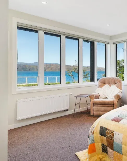 Bright bedroom with large windows overlooking blue water and hills, featuring a chair and quilted bed.
