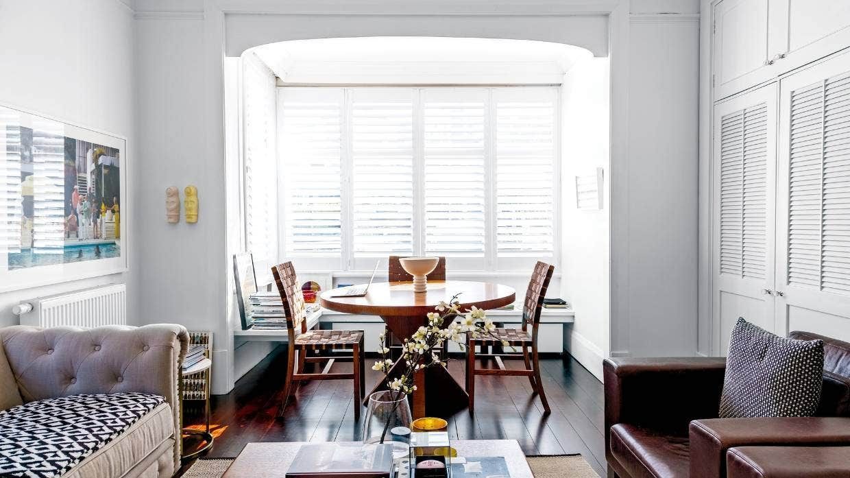 Bright dining room with a wooden table, chairs, sofa, and large white plantation shutters.